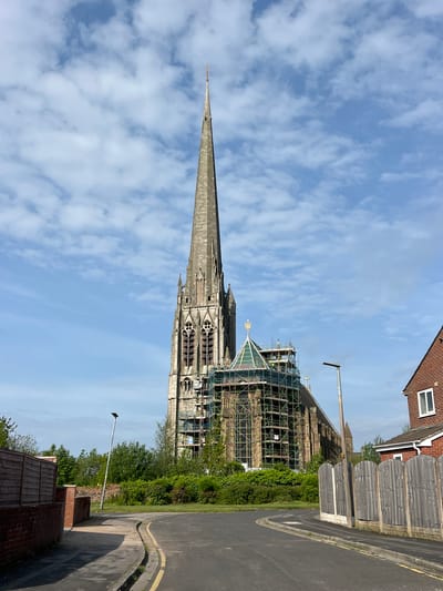 The image depicts a tall, stone church with a prominent spire, partially covered in scaffolding, standing next to a residential street. The church's Gothic architecture is visible in its windows and pointed arches. A street with pavement is located to the right of the church, bordered by fences and foliage. A brick building and wooden fences are seen along the road edge. The sky is blue with scattered white clouds. The location is likely near the city of Preston, United Kingdom.