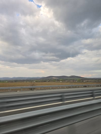 The image displays a wide-angle view of a rural landscape from the perspective of a moving vehicle on a highway in Yazdach, Bulgaria. The foreground is dominated by a multi-tier metallic highway guardrail structure exhibiting motion blur, indicating forward movement. Immediately beyond the guardrail, a two-lane asphalt road with a solid white line on the right shoulder is visible. The midground features an expansive field of dry, light brown vegetation. Scattered across this field are several clusters of deciduous trees, some appearing to be in bloom with white or light green foliage. Further back, a series of rolling hills covered in dark green vegetation rise, forming the distant horizon. The sky above is largely overcast with dense, grey clouds, though small sections of brighter blue sky are visible in the upper right and center-left areas. No people or specific vehicles are depicted within the scene.