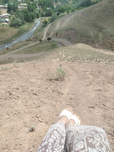 The image captures a scenic view from a hillside overlooking the city of Tashbulak, Uzbekistan. In the foreground, a person is seated on dry, dusty ground, their legs clad in patterned trousers and white sandals. The terrain is sloped and uneven, with sparse vegetation. In the mid-ground, a winding river flows through a valley, bordered by green vegetation and a small settlement of houses. A yellow excavator is visible near the riverbank. The background reveals rolling hills and more distant houses and trees, indicative of a rural or semi-rural landscape. The overall scene suggests a warm, dry climate.