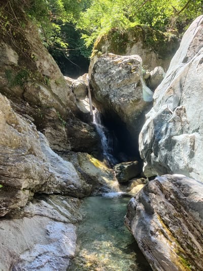 A shallow, clear stream flows over smooth, light-colored rocks, forming small cascades within a rocky gorge. The surrounding rocks are large and grey, with some moss and vegetation growing on them. Trees with green foliage overhang the gorge, and sunlight filters through, illuminating parts of the scene. The water appears cool and inviting, and the environment suggests a natural, secluded outdoor setting. The image is likely taken in a mountainous or forested area. The specific location in the Municipal Unit of Melivia, Greece, is a natural watercourse within a rugged landscape.