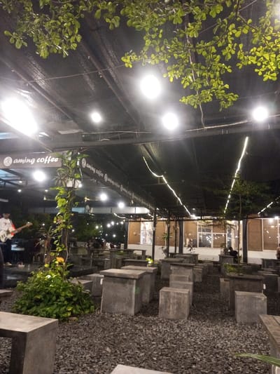 The scene depicts the outdoor seating area of "Aming Coffee," an establishment in Pontianak, Indonesia, at night. A dark, likely mesh or tarp, canopy spans the overhead space, supported by a metal framework. Multiple bright, circular white lights are integrated into this canopy, complemented by linear string lights traversing the area. The ground is covered with dark, loose gravel. Numerous precast concrete blocks, serving as both tables and seating, are arranged throughout the space.
On the far left, a person, partially visible, wearing a white long-sleeved shirt and dark pants, is actively playing an electric guitar, indicating live music. A white keyboard is visible immediately to their right. In the middle-ground and towards the right, several patrons are seated at the concrete tables, their forms indistinct. Green vegetation is interspersed, including hanging plants, ground cover, and small trees, with prominent tree branches and leaves visible at the top of the frame. In the background, a building with illuminated windows and a lighter-colored facade is visible.