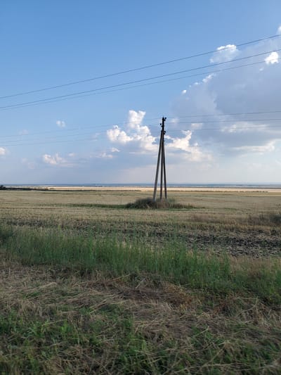 The image shows a wide agricultural field under a partially cloudy blue sky. In the foreground, there is tall green grass and dry, straw-like vegetation. The field itself appears to be harvested, with stubble visible. A wooden utility pole with insulators and wires stands in the middle ground, supporting overhead power lines that stretch across the frame. In the distance, there is a flat horizon line with more fields and a faint appearance of buildings or structures. The scene is devoid of people and any discernable human activity. The overall impression is one of a rural landscape in Moldova, likely near the village of Uyutnoye, given the agricultural setting and the presence of power lines.
