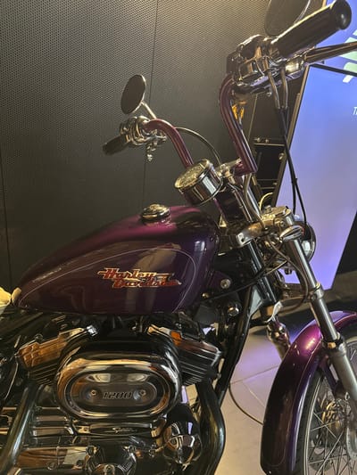 A metallic purple Harley-Davidson Sportster 1200 motorcycle is prominently displayed indoors within the city of Alcobendas, Spain. The motorcycle features high-rise handlebars, a chrome speedometer, and a fuel tank adorned with the "Harley-Davidson" script logo in red and orange. Chrome engine components, including an air cleaner cover embossed with "1200 Sportster," are visible. The motorcycle's front fender and spoked wheel are also metallic purple. The background consists of a dark gray, perforated wall with a grid pattern. To the right, a large vertical screen or display emits a blue and purple ambient light, casting reflections on the floor and motorcycle. No individuals are present in the scene.
