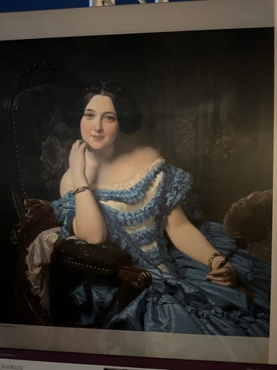 The image is a photograph of a painting depicting a woman seated in an ornate chair, dressed in a voluminous blue gown with ruffled white trim. She has dark hair styled up and is looking towards the viewer with a gentle expression. Her left arm rests on the chair, with her hand near her chin. Her right arm is extended, and she is holding a dark object, possibly a quill or a fan. A bracelet is visible on her left wrist, and another on her right wrist. The background is dark and appears to be a draped fabric or a dimly lit room with decorative elements. At the bottom of the image, text is partially visible, including "MADRAZO" and "ESPAÑOLA", suggesting the painting might be by a Spanish artist named Madrazo. There is no indication of the city of Mazarrón, Spain within the image itself.
