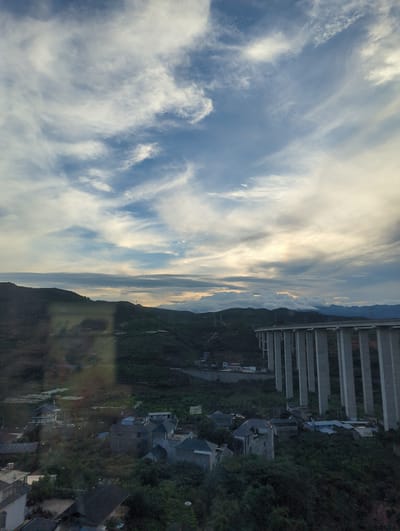 The image captures a landscape near Mengzi City, China, dominated by a vast, elevated bridge with numerous concrete pillars supporting a roadway. The bridge stretches across a valley filled with lush green vegetation and a scattering of residential buildings. The sky above is a dramatic mix of blue and white clouds, with some areas appearing bright and others shadowed, suggesting either sunrise or sunset. The terrain is hilly, with layered mountains in the distance. A reflection is visible on the surface from which the image was taken, obscuring some of the foreground details of the town and vegetation. Several houses, some with tiled roofs, are nestled amongst the dense greenery. Electrical towers can be seen on the hillsides.