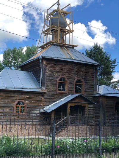 The image shows a rustic wooden building, likely a church or chapel, in Izdeshkovo, Russia. The structure is made of horizontal logs and features a gray metal roof. There is scaffolding around the upper part of the building, suggesting ongoing construction or renovation. The building has several arched windows with wooden frames. A covered entrance with a porch and a dark metal gate is visible. The scene is set against a backdrop of trees and a blue sky with white clouds. A black wrought-iron fence with decorative elements encloses the property, and a flower bed with white and pink flowers is visible in front of the fence. Power lines are present in the upper portion of the image.