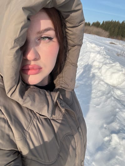 The image depicts a young woman in a snowy outdoor environment, identified as Pavlovka, Russia. The woman is positioned from the chest up, angled slightly to the viewer's left, with her gaze directed at the camera. She has reddish-brown hair, visible around her face, green eyes accentuated with dark eyeliner, and full lips colored with pink lipstick. She is wearing a light brown or taupe quilted puffer jacket with the hood drawn up, partially shadowing the left side of her face. A black collar or garment is visible beneath the jacket's opening. Strong natural light illuminates the scene, creating shadows on her face and clothing. The background consists of a vast expanse of white snow, showing some irregularities and potential tracks. To the right, the snow is visibly mounded. In the mid-ground, a line of leafless trees is visible, followed by a dense coniferous forest under a clear, bright blue sky.
