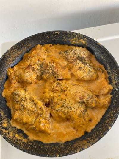 This media file shows a close-up, overhead view of chicken in a creamy sauce within a black frying pan. The pan is resting on a white surface, possibly a countertop, with some light casting shadows on the left. The chicken pieces are coated in a thick, orange-hued sauce, and appear to be seasoned with herbs, likely dill. The sauce has a slightly bubbly texture, suggesting it is either being cooked or has recently been cooked. The pan itself has a speckled non-stick surface, and some sauce has splattered onto the rim and onto the white surface surrounding the pan. The overall impression is of a home-cooked meal being prepared or served. There are no people, animals, or text visible in the image. The lighting appears to be artificial, as there are no strong natural light cues. The scene is domestic and focused entirely on the food.