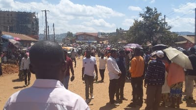 A crowd gathers on a dusty road, with buildings under construction visible in the background. People are dressed in a variety of casual clothing, with some using umbrellas for shade. Vehicles and more people are visible in the distance, suggesting a busy town center. The foreground is dominated by a man with a light pink shirt, observed from the back. The overall impression is a bustling scene of everyday life in a developing area.