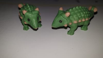 Two green toy dinosaurs with light pink accents are positioned on a white, textured surface. The dinosaur on the left is facing left, and the dinosaur on the right is facing right, with their heads turned towards each other. Both dinosaurs have black eyes and appear to be Ankylosaurus figures. The background is dark and out of focus. The image was taken in Vrchotovy Janovice, Czechia.