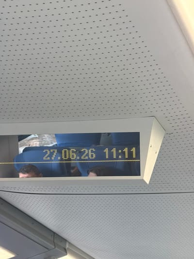 The image shows a view from inside a train looking upwards towards a digital display. The display shows the date "27.06.26" and the time "11:11" in yellow LED dot matrix characters against a dark background. Below the display, the heads of two passengers are visible, partially obscured by the display unit. They are seated in blue train seats. The ceiling of the train carriage is visible, covered with a white perforated material. The overall environment suggests the interior of a public transport vehicle, likely a train, given the presence of seats and the digital display showing time and date. There are no obvious indicators of the specific city of Ушаки, Russia within the image itself.