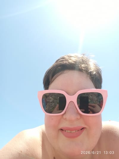 In this close-up, over-the-shoulder shot, a person wearing pink, oversized sunglasses is captured against a bright, cloudless blue sky. The sunglasses' lenses reflect a scene of pebbles and a hand holding a dark-colored smartphone. The person's face is partially visible, showing their mouth with pink lipstick and their upper chest and shoulder. The sun is a bright, blown-out white light source in the upper part of the frame, creating lens flare. A timestamp in the lower right corner reads "2026/6/21 13:03". The scene appears to be outdoors, likely in a sunny, potentially coastal location. Given the context of Bolshoy Sochi, Russia, it's plausible this photo was taken on one of its many beaches, with the pebbles in the sunglasses reflecting the beach environment and the clear sky suggesting a summer day.