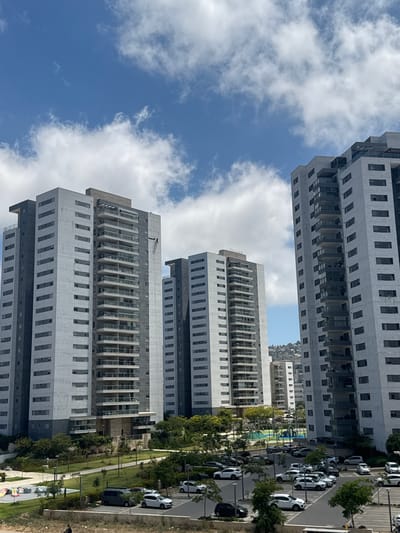This media file captures a view of modern residential buildings in Tirat HaCarmel, Israel. The scene is dominated by several tall, multi-story buildings with repetitive window patterns and balconies. The buildings are constructed with a mix of light-colored concrete or stone cladding and darker elements.
In the foreground, on the right side of the image, is a parking lot with several white cars and some darker vehicles parked in a row. A sidewalk or path runs alongside the parking lot, bordered by green grass and trees. Further into the background, more buildings are visible, some with a mountainous or hilly landscape behind them. A playground area with colorful structures is also visible between some of the buildings.
The sky is a bright blue with scattered white clouds, suggesting it is daytime, likely mid-morning or afternoon. The lighting indicates a sunny day. The overall impression is one of a contemporary urban or suburban environment, characterized by modern architecture and residential development. There is no visible text in the image. The scene appears peaceful and ordinary, depicting a typical day in a residential area.