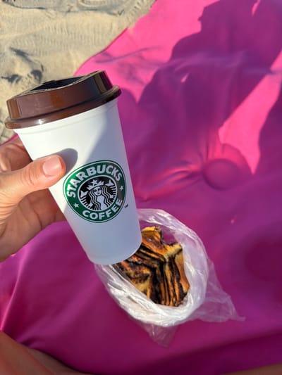 In Urbanització Cales I Talaies, Spain, a person's hand, with neatly manicured fingernails, holds a white Starbucks coffee cup. The cup features the iconic green Starbucks logo. The cup is positioned over a plastic bag containing a slice of chocolate babka. The scene is set against a bright pink fabric, likely a beach towel or blanket, with sand visible in the upper left corner. Shadows, cast by an unseen object or person, create a dramatic effect on the pink fabric. The lighting suggests it is a sunny day, likely outdoors.