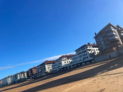 The image displays a beachfront scene in Zarautz, Spain, captured with an upward and rightward tilted perspective. The foreground consists of a wide expanse of light brown, compacted sand, traversed by elongated dark shadows extending diagonally from the lower-left towards the upper-right, indicating a low sun angle and direct illumination. No individuals, animals, or distinct objects are visible on the sandy surface.

In the midground, a continuous row of multi-story buildings borders the edge of the beach, likely fronting a promenade or street. These structures vary in architectural style, typically ranging from three to six floors, and feature balconies, numerous windows, and diverse facade materials including light-colored stucco, patterned brick, and dark panels. Several ground-level sections exhibit large glass fronts, suggesting commercial establishments. The building on the far right has a distinct, darker brick-like facade with multiple visible floors and windows.

The background is dominated by a clear, vibrant blue sky. A single, faint white contrail from an aircraft is discernible in the upper-left quadrant of the sky, and a few thin, wispy white cloud formations are present near the horizon line on the left side of the frame.