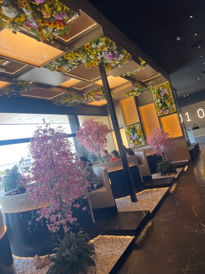 An interior view shows a multi-tiered decorative wall structure, featuring alternating yellow-lit compartments and boxes filled with mixed floral arrangements dominated by sunflowers. Adjacent to these, faux cherry blossom trees stand in white pots, reaching toward a skylight. Tables and chairs are situated in the lower part of the frame, occupied by individuals, some wearing masks. The setting appears to be an upscale restaurant or commercial space in Torreforta, Spain.