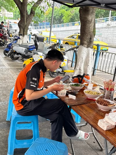 The scene is an outdoor street setting, possibly a temporary eatery, within the 龙溪街道 region of China.

A young man, wearing an orange and black shirt, is seated on a blue plastic stool at a wooden table. He is looking at and holding a smartphone in his hands. On the table in front of him are various bowls of food, chopsticks, and wrapped food items.

Behind him, multiple motorcycles and scooters are parked along the street. In the background, a row of taxis is visible, along with a sidewalk and trees. A tree trunk appears to be close to the table, its lower part painted white. A structure, possibly part of a temporary tent or covering, is visible on the left side of the image.