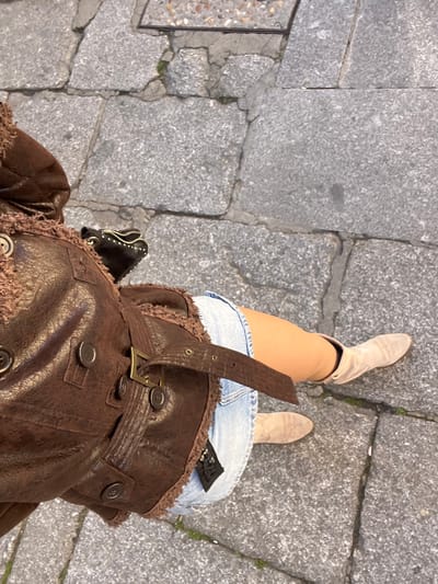 The image shows a downward-angled view of a person standing on a grey stone-paved area in Salamanca, Spain. The paving stones are irregular in shape and size with narrow joints. The person is wearing light beige cowboy boots, a denim skirt, and a brown leather-like jacket with a fur-like trim and a buckled belt. A small black bag with studs is visible on their side.