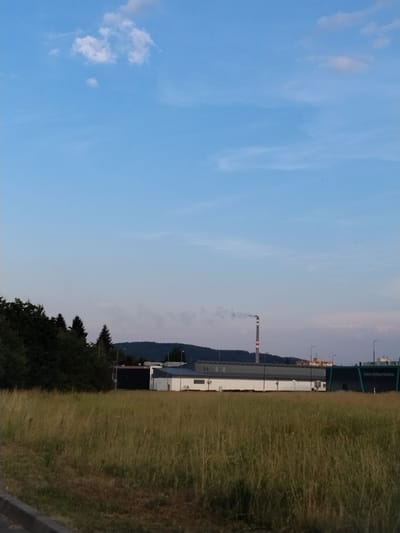 The image depicts an industrial landscape with a clear blue sky and some wispy clouds. A tall, red and white striped smokestack emits a plume of smoke on the right side of the frame, suggesting an active industrial operation. In the background, a long, white building with a dark roof runs horizontally, with what appears to be a darker industrial structure at its far end. Streetlights are visible along the building. A dark, tree-covered hill or mountain is situated behind the buildings. To the right of the buildings and smokestack, a dense area of tall, dry grass fills the foreground, gradually turning into a darker, more lush green area of trees and foliage further to the right. A faint, dark horizontal line runs along the bottom right edge of the image, possibly a road or pathway. The lighting suggests it is either early morning or late afternoon. The scene is located in Strakonice, Czechia. Text is visible on a dark banner above the white building; it reads "Třebíčský nábytek pro Váš domov".