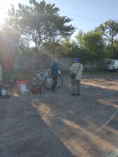 Two men are standing in an outdoor lot next to a concrete fence. The man on the left is wearing a blue jacket and a cap, and is looking to the right. The man on the right is wearing a yellow and black striped shirt, camouflage pants, and a blue cap, and is looking to the left. In front of the man on the left are two bicycles, one is partially disassembled. To the left of the bicycles are boxes and coils of yellow cable. To the right of the bicycles is a bucket and a bicycle pump. In the background, a white van is parked next to the fence. The ground is dirt and gravel. The scene is illuminated by bright sunlight. The location is the city of Irshansk, Ukraine.