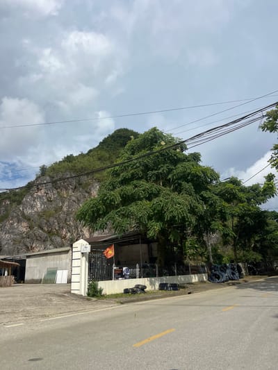 The image shows a scene in Quản Bạ Commune, Vietnam, characterized by a steep, rocky mountain face covered in vegetation. In the foreground, a paved road runs horizontally, with yellow dividing lines. To the left of the road, there is a building with a concrete wall and a section that appears to be a workshop or storage area, with some glass panes leaning against the wall. Adjacent to this building is a white pillar with black stripes, bearing a sign that reads "Hạn Coffee & Tea." A black fence encloses a small area behind the pillar, where a Vietnamese flag is flying. Several large trees with lush green foliage are prominent in the middle ground, obscuring parts of the mountain. To the right of the fence, a pile of black coiled pipes or hoses is stacked on the ground, and more are visible further along the roadside. Overhead, a network of power lines crisscrosses the sky, which is partly cloudy. The overall environment suggests a semi-rural or commercial area bordering a natural landscape. No people are visible in the image.