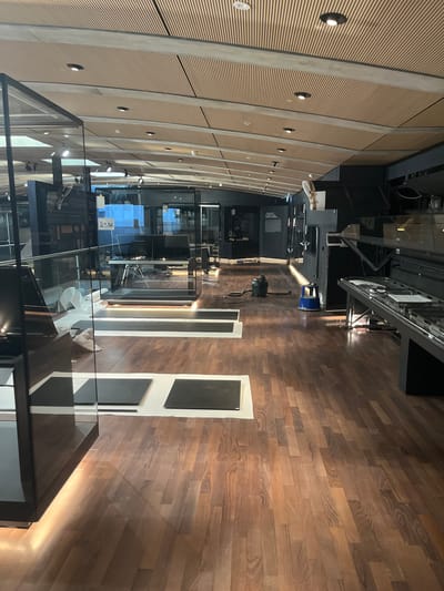The image depicts the interior of a museum exhibition space in Cologny, Switzerland. The room features a polished wooden floor and a ceiling with a slatted wood finish. Various display cases made of glass and dark metal are present throughout the space, some containing exhibits or equipment. In the mid-ground, a vacuum cleaner and a small blue stool are visible on the floor, suggesting ongoing cleaning or setup activities. The lighting appears to be a mix of overhead fixtures and accent lighting along the display cases. The overall impression is one of a modern, well-lit exhibition area undergoing preparation or maintenance.