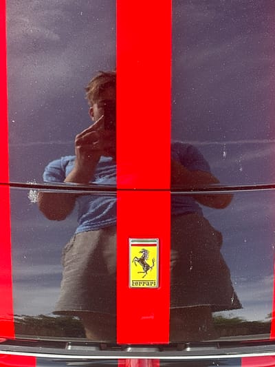 This is a close-up image of the rear of a Ferrari, likely a model with a painted black rear section and a horizontal red stripe. The iconic Ferrari badge is visible on the right side of the car. The surface of the car reflects the environment, including a person taking a photograph, and suggests the presence of a wet surface. The location is Jestetten, Germany. The reflection of the person suggests they are standing close to the vehicle, likely taking a picture of the Ferrari. The overall environment appears to be outdoors, possibly at a car show or event.