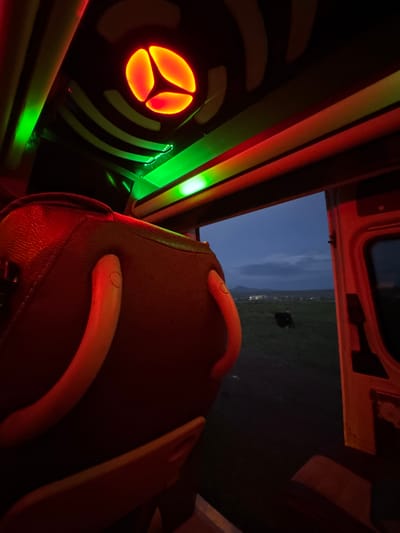 The image depicts the interior of a vehicle, likely a van or bus, with customized lighting. A circular orange light fixture with a three-lobed design is mounted on the wall. Green LED strip lights are visible as well.
A vehicle seat with a reddish-brown leather or vinyl covering is in the foreground. Two light-colored plastic or composite grab handles are attached to the seat.
Through the open rear doors of the vehicle, a twilight or nighttime landscape is visible. A dark, indistinct shape, possibly an animal, is visible in the dark field. Distant lights, suggesting a settlement, are visible on the horizon. The city of Anipemza, Armenia is not identifiable in the image.