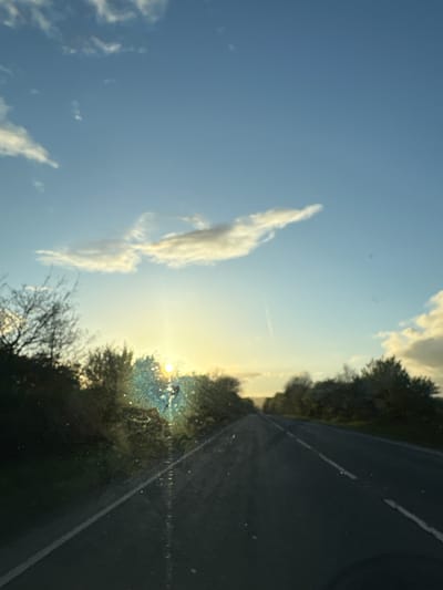 The image depicts a view from inside a vehicle, possibly a car, looking out onto a rural scene. The sky is blue with some scattered white clouds and a contrail from an airplane. The sun is visible near the horizon, obscured by trees. A road with a white line on it is seen on the right side of the image. The location is near Oswestry, United Kingdom, based on geographical context. No people are visible in the frame.