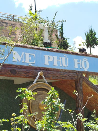 The image depicts a sign with white lettering on a light blue background, which reads "ME PHÙ HO". The sign is mounted on a wooden beam structure that appears to be part of an entrance or gateway. Below the sign, a large, round, bronze-colored gong is suspended by a thick rope with a golden knob. In the foreground, several leafy green plants with small buds are visible, partially obscuring the gong. In the background, a white statue of an angelic figure holding a staff stands on a dark base, positioned behind the sign and to the left of the frame. Further back, there is a stone wall with a railing and greenery, including trees, one of which has solar panels attached to a pole nearby. The sky is blue with scattered white clouds. This scene is located in K'Rèn, Vietnam.