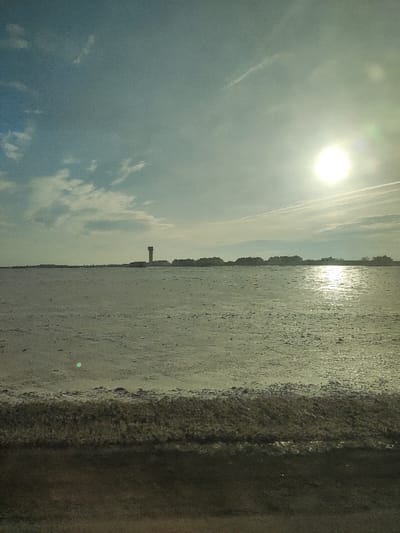 The image captures an expansive, somewhat stark outdoor landscape, likely taken during bright daylight, either early morning or late afternoon, given the low angle and intensity of the sun. The scene is dominated by a vast, flat expanse under a partly cloudy sky.
The sky occupies roughly the top half of the image, displaying shades of light blue with scattered white and grey clouds. On the right side of the sky, the sun is a brilliant, overexposed white orb, casting a strong glare and lens flare across that portion of the image. Faint, linear cloud formations or contrails are visible horizontally across the upper sky, particularly on the right.
Below the sky, the middle ground is a broad, flat area that appears to be a body of water, possibly a large lake or an estuary, or perhaps a flat, open field that is damp or covered in very short vegetation. It has a muted, greenish-grey hue and a textured surface. On the right side of this expanse, the sunlight reflects intensely, creating a shimmering path of light on the surface.
Along the distant horizon, a line of dark, low-lying structures is visible, silhouetted against the brighter sky. These appear to be buildings, possibly houses or commercial properties. A distinctive tall, slender tower, dark brown or grey in color, stands out on the left side of the distant horizon line, rising higher than the other structures. It could be a water tower, a lighthouse, or a communication tower.
The foreground of the image features a darker, textured strip that occupies the bottom portion. This area appears to be a closer view of the immediate ground, possibly a sandy or muddy shoreline, or perhaps the edge of a road or embankment from where the photo was taken. Its rough texture and darker tones contrast with the lighter expanse beyond. There are no people, animals, or visible text in the image. The overall impression is one of wide-open space and calm, with the sun being a prominent focal point.