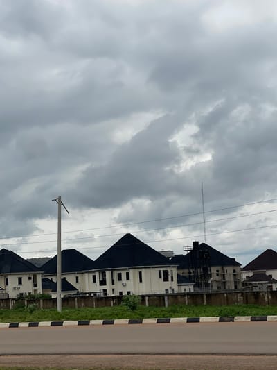 In Immigration Quarters, Nigeria, a residential area features a row of modern, multi-story houses with dark, pitched roofs and light-colored facades. The houses have numerous windows and visible air conditioning units. A concrete wall, topped with barbed wire, separates the properties from a grassy area. In the foreground, a road with a black and white striped curb runs parallel to the houses. A tall utility pole with wires extends upward in the middle ground, and a communication tower is visible in the distance. The sky is overcast with heavy, gray clouds, suggesting a cloudy or potentially rainy day. There are no people or vehicles visible in the scene.