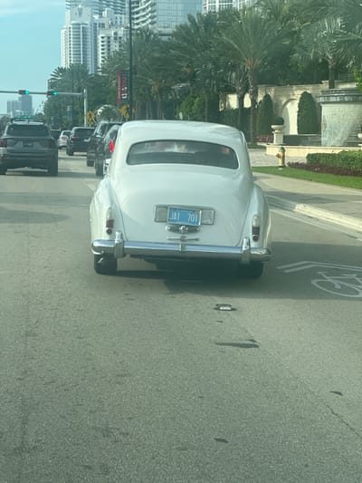The image captures a rear view of a white vintage car, possibly a Rolls-Royce, driving on a sunny day in Sunny Isles Beach, Florida, United States of America. The car has a license plate that reads "JAI 701" and is labeled as "FLORIDA ANTIQUE". In the background, modern high-rise buildings and palm trees are visible, characteristic of a beachfront city. Traffic is moderate, with other vehicles, including SUVs, also on the road. The environment appears to be a well-maintained urban area with a paved road and a sidewalk.
