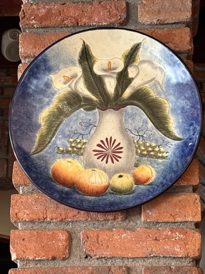 A decorative ceramic plate depicting a still life of calla lilies, grapes, and oranges is mounted on a brick wall. The plate is circular with a dark blue rim. The central image features a white vase with a red asterisk-like design, from which large, white calla lilies with green leaves emerge. Bunches of green grapes and several oranges are arranged at the base of the vase. The background of the plate is a mottled blue. The plate is positioned on a section of exposed brick wall, with some mortar visible between the bricks. The brickwork appears rustic and aged, common in traditional Mexican architecture, potentially in a restaurant or a home in Tepeji de Ocampo, Mexico, known for its artisan crafts. A small, white, circular object, possibly a security sensor, is visible on the brick wall to the left of the plate.