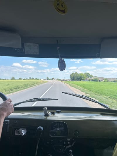 The media file captures an interior view from the driver's perspective of a vehicle, likely a truck, driving on a road. The scene is set in a rural area with green fields and scattered trees lining both sides of the asphalt road, which stretches into the distance. The sky is bright blue with fluffy white clouds, suggesting fair weather and daytime.
Inside the vehicle, the dashboard is visible with various instruments, including a speedometer. A hand is seen gripping the steering wheel, indicating the vehicle is in motion. On the windshield, two windshield wipers are present. Hanging from the interior of the vehicle, near the windshield, are a yellow smiley face sticker and a blue oval-shaped object with some text on it. On the left side of the interior, a grey sun visor is visible, and a sticker with diagrams and text is affixed to the inner wall. The overall impression is of a journey through the Belarusian countryside on a sunny day.