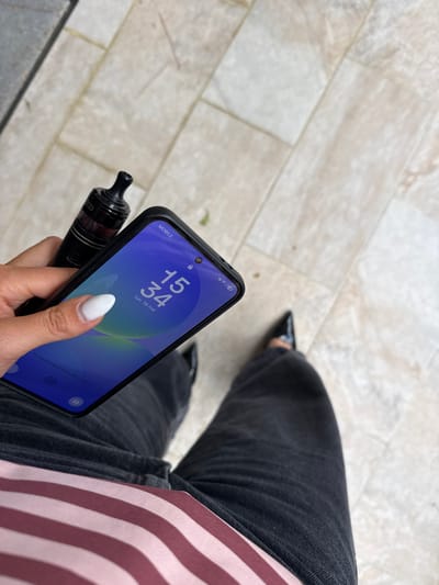 A person is holding a smartphone and a vape device while sitting on a light-colored tiled surface. The phone displays a lock screen with the time 15:34 and the date Lun 18 Mai. The person is wearing dark jeans and a pink and white striped shirt, with shiny black shoes visible in the background. The ground is composed of light gray tiles arranged in a linear pattern. The city or town is identified as Les Arcs, France.