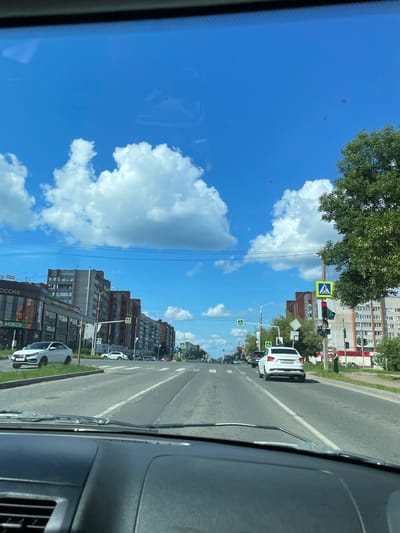The image captures a view from inside a vehicle driving on a sunny day in Kirovo-Chepetsk, Russia. The scene depicts a city street with multi-story apartment buildings lining both sides. The road has multiple lanes and is marked with white lines. Pedestrian crossing signs are visible at intersections. Several cars are driving on the road, and some are parked. A large green tree is on the right side of the image. The sky is bright blue with scattered white clouds.