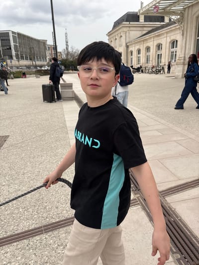 A young male, appearing to be a pre-teen, stands in the foreground, centered slightly to the left, looking directly at the camera with a neutral expression. He has short, dark hair and wears clear-framed glasses. His attire consists of a black short-sleeved t-shirt with the word "LABRAND" (or similar) printed in light blue on the chest, accompanied by a vertical light blue stripe on the shirt's right side. He wears light beige trousers. His left hand holds a dark, textured rope or leash extending out of frame to the left.

The scene unfolds on a paved public plaza featuring light-colored stone tiles and dark linear grates. In the background, to the right, is the historic Blois train station (Gare de Blois) in Blois, France. This building has a light-colored stone facade with multiple arched windows on the ground floor and a dark mansard roof. A modern glass and metal canopy with the "SNCF" logo extends from its entrance. Bicycle racks with parked bicycles are visible near the station.

To the far left of the frame, a modern building with a facade composed of horizontal gray slats is visible. Several individuals are dispersed across the plaza: one person in dark clothing with a black rolling suitcase stands near a low concrete barrier to the mid-left, another partially obscured person with a dark backpack is behind the main subject, and a person in a dark blue jumpsuit walks away to the right. The sky is overcast. A tall, dark pole stands in the mid-ground, and a distant communication tower is visible on the horizon.