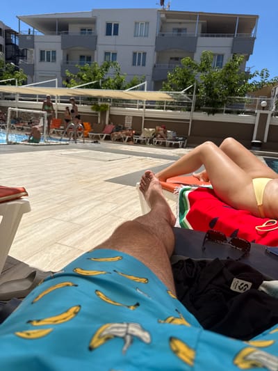 The image depicts a poolside scene on a sunny day in Ayvalık, Turkey. In the foreground, a person's legs are visible, wearing blue swim trunks with a banana print. Another person's legs, wearing a yellow bikini bottom, are also visible on a lounge chair adorned with a red and green striped towel and a watermelon pattern. Sunglasses and a black t-shirt are placed nearby on the lounge chair. In the background, a swimming pool is visible with people lounging on chairs and swimming. A multi-story building with balconies and trees surrounds the pool area, typical of a resort or hotel in Ayvalık. The ground around the pool is paved with light-colored tiles.