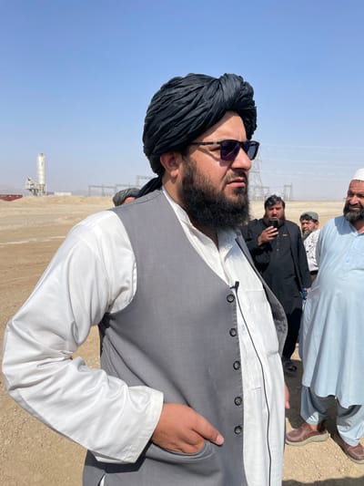 An outdoor scene in Pir Dust, Afghanistan, depicts a group of men on arid, light-brown ground under a clear blue sky. In the foreground, a man with a dark beard and mustache, wearing a black turban, dark sunglasses, a white tunic, and a grey waistcoat, stands with his right hand in his pocket. A lavalier microphone is clipped to his white tunic. He looks towards the right. In the midground to his right, another man in dark traditional clothing holds a smartphone, seemingly recording. Further right, a third man in a light blue tunic and white cap is visible. The background features industrial structures, including a tall, light-colored silo and metal electricity pylons, against a hazy, dusty horizon.