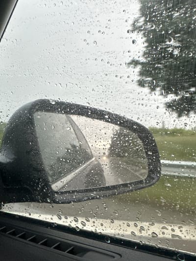 The image provides a close-up view from inside a vehicle, looking out through a window heavily covered in numerous water droplets, indicating rain. The droplets vary in size, from tiny specks to larger, more defined spheres, distorting the view beyond.

A prominent black car side mirror is positioned in the middle-right of the frame, also dotted with raindrops. The mirror reflects a blurred scene from behind the vehicle, showing indistinct light sources or shapes, possibly hinting at a road or other vehicle features.

Through the wet window, the background is highly blurred, with dark, shadowy forms of trees visible on the left side. The right side of the background appears lighter, suggesting an overcast sky or a light-colored structure like a building or pole. The overall lighting is diffused, consistent with a cloudy, rainy day.

No people or explicit activities are visible, but the scene suggests being stationary or moving slowly in a vehicle during a rain shower. There is no visible text in the image. The setting is a rainy day, viewed from within a car, in Zvezditsa, Bulgaria.