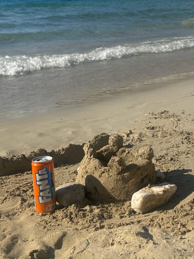 An orange Fanta can rests upright in the sand on a beach. The can is partially obscured by a small sandcastle. Several rocks are scattered around the sandcastle and the can. In the background, the blue ocean meets the sandy shore with gentle waves. The scene appears to be in Agios Nikolaos Municipal Unit, Greece.
