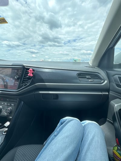 The image, captured from inside a vehicle, shows a dashboard with a pink figurine clipped to an air vent on the right side. The dashboard is dark-colored and features a central infotainment screen displaying navigation, air conditioning controls, and buttons on the left. The viewer's legs, clad in light blue jeans, are visible in the lower portion of the frame. Outside the car, a cloudy sky is visible through the windshield. The image is framed to suggest a person is seated in the driver's or passenger's seat. The scene appears to be an outdoor setting, possibly a road or parking area, given the view of the sky and distant elements. The location is identified as Manolsko Konare, Bulgaria.