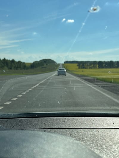 The image shows a view from a car window looking out at a road. The perspective is from the passenger seat, looking forward and slightly to the right. The road is paved and appears to be a highway or a main road, with clear lane markings. There are dashed white lines on the right side of the road, indicating the edge. In the distance, a single car, possibly a dark-colored SUV or van, is traveling in the same direction.

To the left of the road, there is a grassy field with scattered trees. The sky is bright blue with some scattered clouds, suggesting it is daytime, likely late morning or afternoon. The sun appears to be shining, casting a bright light on the scene.

The window itself has some imperfections, including a few small white spots or smudges, and what appears to be a small chip or impact mark with a circular ripple effect on the left side of the frame. The right edge of the image shows a textured gray surface, likely part of the car's interior, possibly the door panel or dashboard.

The overall impression is one of a straightforward drive on a clear day in a rural or semi-rural setting in Russia, specifically Ulukulevo. There are no people clearly visible, and the focus is on the road and the surrounding landscape. The mood is neutral, conveying a sense of travel and observation.