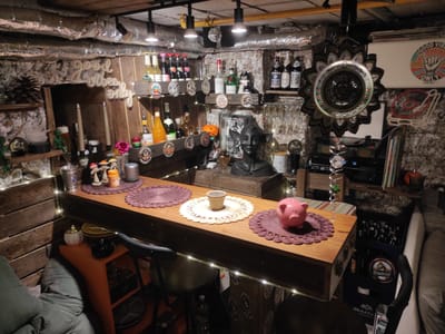 The image depicts a dimly lit, rustic indoor bar area, possibly a basement or man cave. The walls are a rough, textured stone, and the ceiling is covered in exposed, silver-wrapped insulation and pipes, with several black spotlights providing illumination. Wooden planks line some of the walls and form shelves. A central wooden bar counter runs horizontally, laden with various items.
On the bar, to the left, are two doilies, a small metal cup, a beige ceramic cup, and another doily. To the right of these is a pink piggy bank. Behind the bar, on shelves, are numerous bottles of alcohol, including wine and spirits, some with decorative labels. There are also glasses hanging from a rack. In the center background, a dark bust of a person with elaborate headwear stands on a shelf. Various decorative objects are scattered throughout, including small mushroom figures, candles, a small orange pumpkin, and a glass sphere. String lights are draped along the front of the bar, adding to the ambiance.
To the left of the bar, a chair with a dark seat and back is pulled up. In the foreground to the left, a large, plush grey cushion is visible. To the right of the bar, crates of beer bottles are stacked, and a part of a couch or seating area is visible. A large, circular, mandala-like decoration hangs on the wall to the right, and a "Good Vibes Only" poster is also displayed. A turntable and audio equipment are visible on a shelf in the background.
The scene suggests a personal bar setup, likely in a private residence. Without additional information or visible landmarks, it is not possible to definitively locate this scene within the city of Waldbronn, Germany.