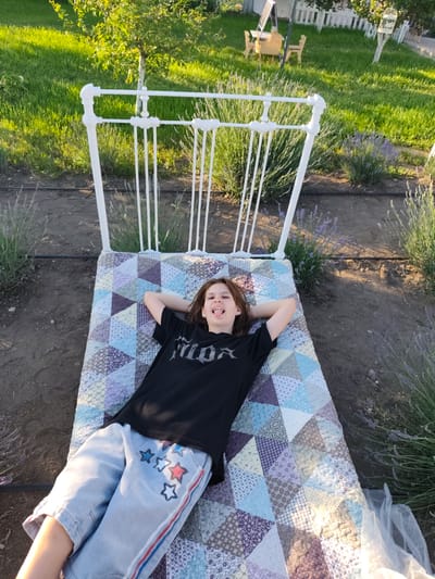A young person with brown hair lies on their back on a quilted mattress on the ground. The person is wearing a black t-shirt with a graphic print and light blue shorts with red and white star designs and red and blue stripes along the sides. Their arms are behind their head, and their tongue is sticking out. The mattress is placed on the ground, possibly in a garden or park setting, with a white metal bed frame behind it. In the background, there is green grass, lavender bushes, and a white picket fence. The scene appears to be outdoors during daylight. The location is identified as Nechaiane, Ukraine.