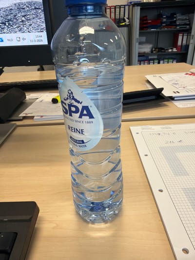 A clear, partially-filled plastic bottle of SPA Reine Natural Mineral Water, featuring a blue screw cap and a white label, stands upright on a light-colored wooden desk. The label displays the brand "SPA" with a figure holding two buckets, and the text "REINE NATURAL MINERAL WATER PROTECTED SINCE 1889".

To the left of the bottle, a black computer monitor displays a desktop background of a rocky surface. The monitor's lower-left corner shows "NLD", a network icon, a speaker icon, and the date "11-3-2026" and time "15:44". A black computer keyboard is partially visible in the lower-left foreground. Various papers are scattered on the desk, including one with a yellow highlighter resting on it, and another open notebook on the right with handwritten entries "1359,54 €" and "276 €" on a grid pattern. A black folder is also present on the desk.

The background consists of office furniture, specifically a metal shelving unit containing numerous binders and files in various colors (red, white, blue, black). No individuals are visible, and no active interactions are depicted. The scene is an interior office environment located in Apeldoorn, Netherlands.