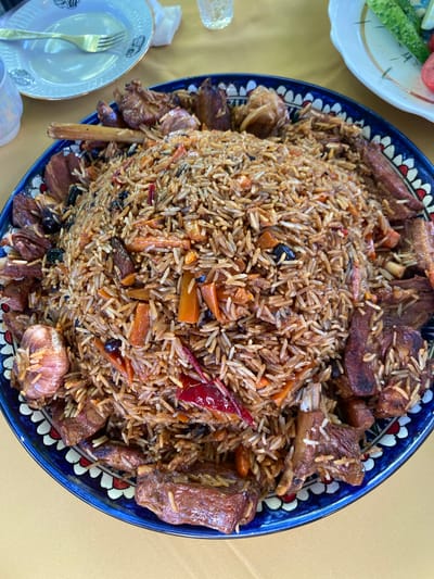 A top-down, close-up shot showcases a large, circular blue and white platter filled with pilaf. The pilaf is a mound of long-grain rice, interspersed with chunks of meat, carrots, and what appear to be dried fruits or raisins. Several whole garlic bulbs and dried red chili peppers are also visible within the rice. The meat pieces are varied in size and shape, some appearing to be ribs with bone fragments. The platter is ornately decorated with a geometric pattern in blue, red, and white around its rim.
In the upper left of the frame, a white plate with a gold rim and a fork rests on a yellow tablecloth. To the upper right, another white plate holds sliced cucumbers and tomatoes. The background is dominated by the yellow tablecloth.
The image is focused on the pilaf, with elements in the background slightly blurred. The lighting is bright, suggesting an outdoor or well-lit indoor setting. No people are visible in the image. The scene appears to be a meal setting, possibly a traditional dish being served. The location is specified as Znamenski Sel'ski Savet, Belarus, though there are no specific visual cues in the image to confirm this location.