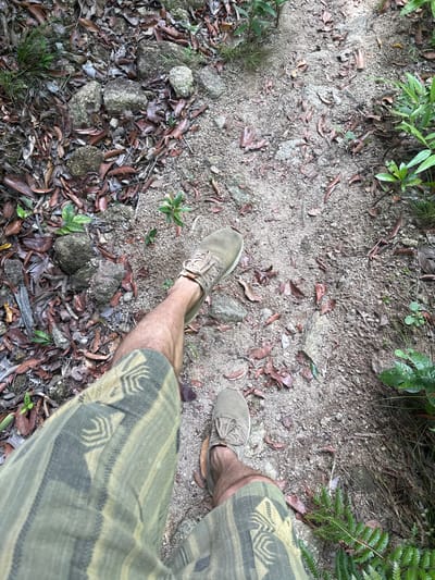 The image depicts a first-person perspective of a person standing on a dirt path in Moo 7, Thailand. The path is covered in loose dirt, rocks, and fallen leaves. The person is wearing khaki-colored sneakers and patterned khaki shorts. Vegetation borders the path on both sides.