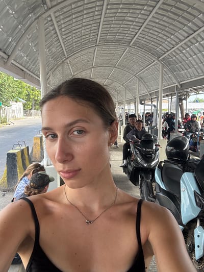 In Upt Tambak Sari, Indonesia, a young woman with brown hair pulled back and wearing a black tank top takes a selfie under a covered walkway. Her face is turned towards the camera, with a necklace visible on her chest. Behind her, a line of motorcycles and people waiting is visible. The walkway has a corrugated metal roof supported by white pillars, and in the background, trees and buildings can be seen. A road with yellow and black striped barriers is also present. Some individuals on motorcycles are wearing helmets and casual clothing.
